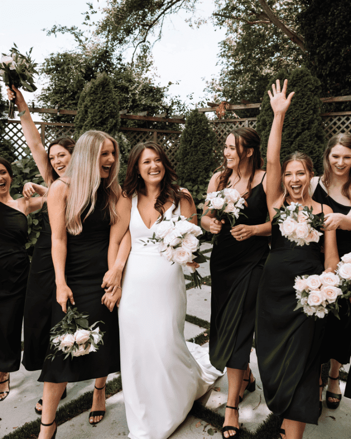 Bride and bridesmaids in black dresses smile and celebrate outdoors, holding bouquets of flowers.